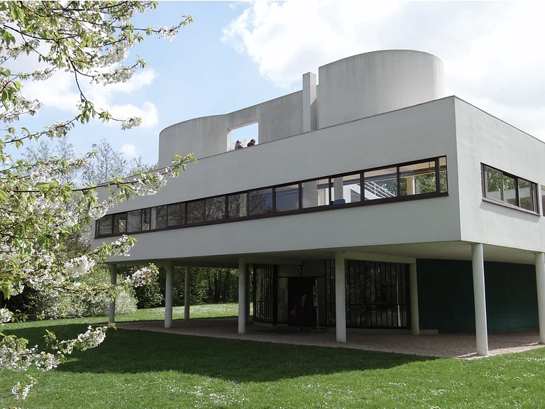 Villa Savoye by Le Corbusier, a white modernist house raised on pilotis with ribbon windows and a rooftop terrace surrounded by green lawn and blossoming trees.